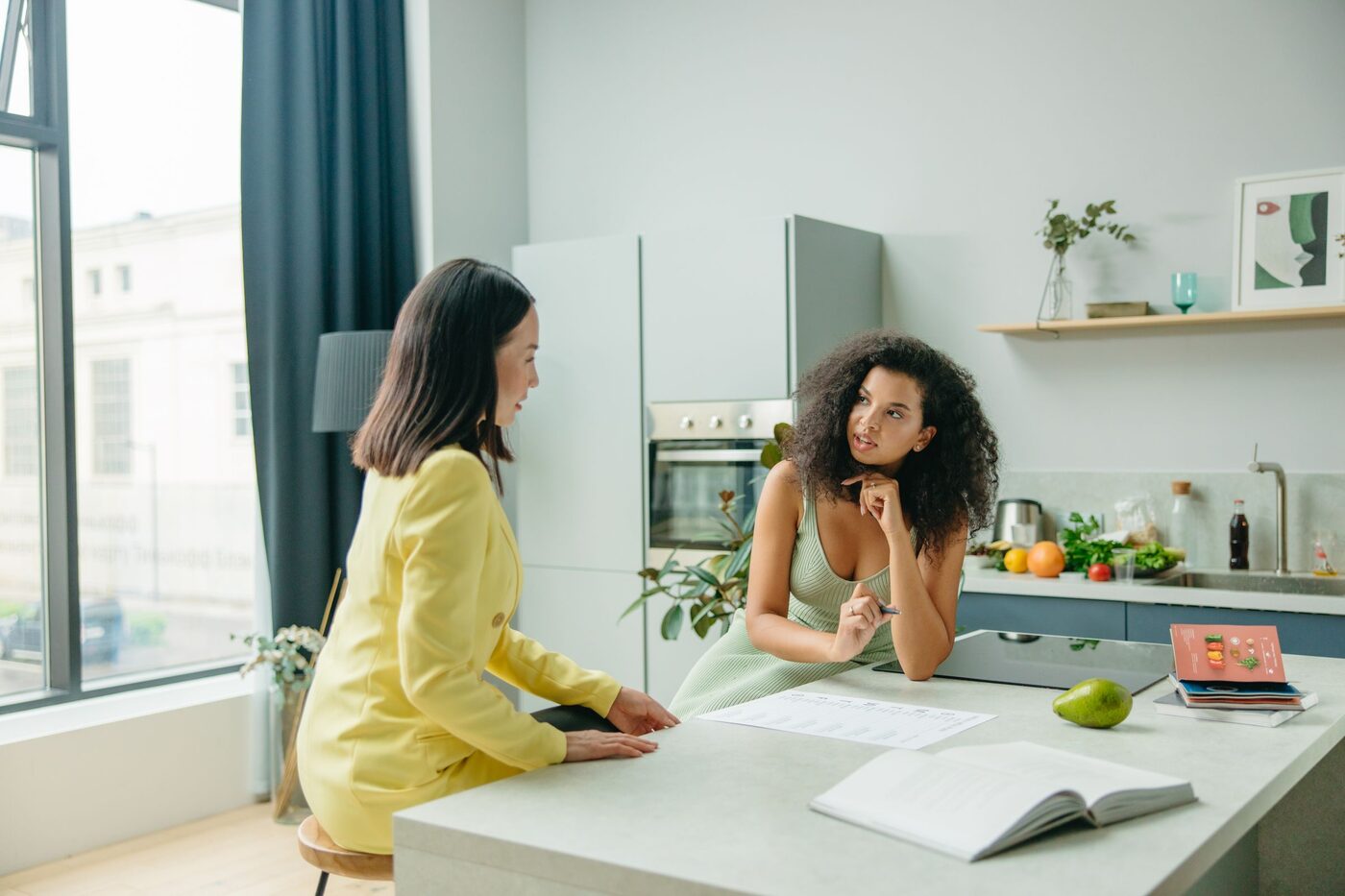 Two women talking across a sunlit kitchen counter, notes and fresh fruit between them.