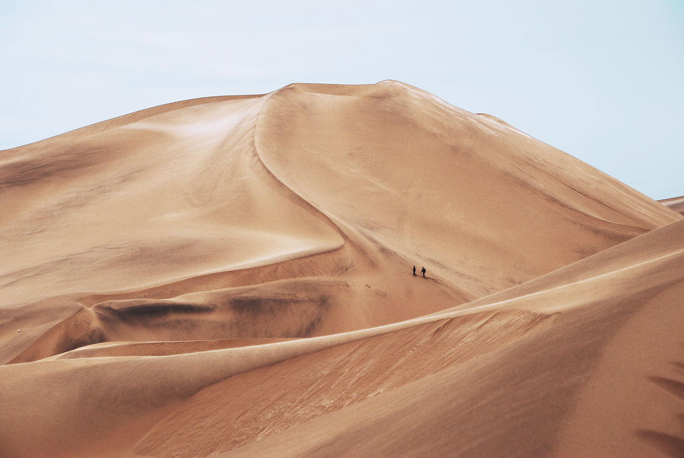 Two people walking together along the ridge of a vast sand dune, dwarfed by its scale.