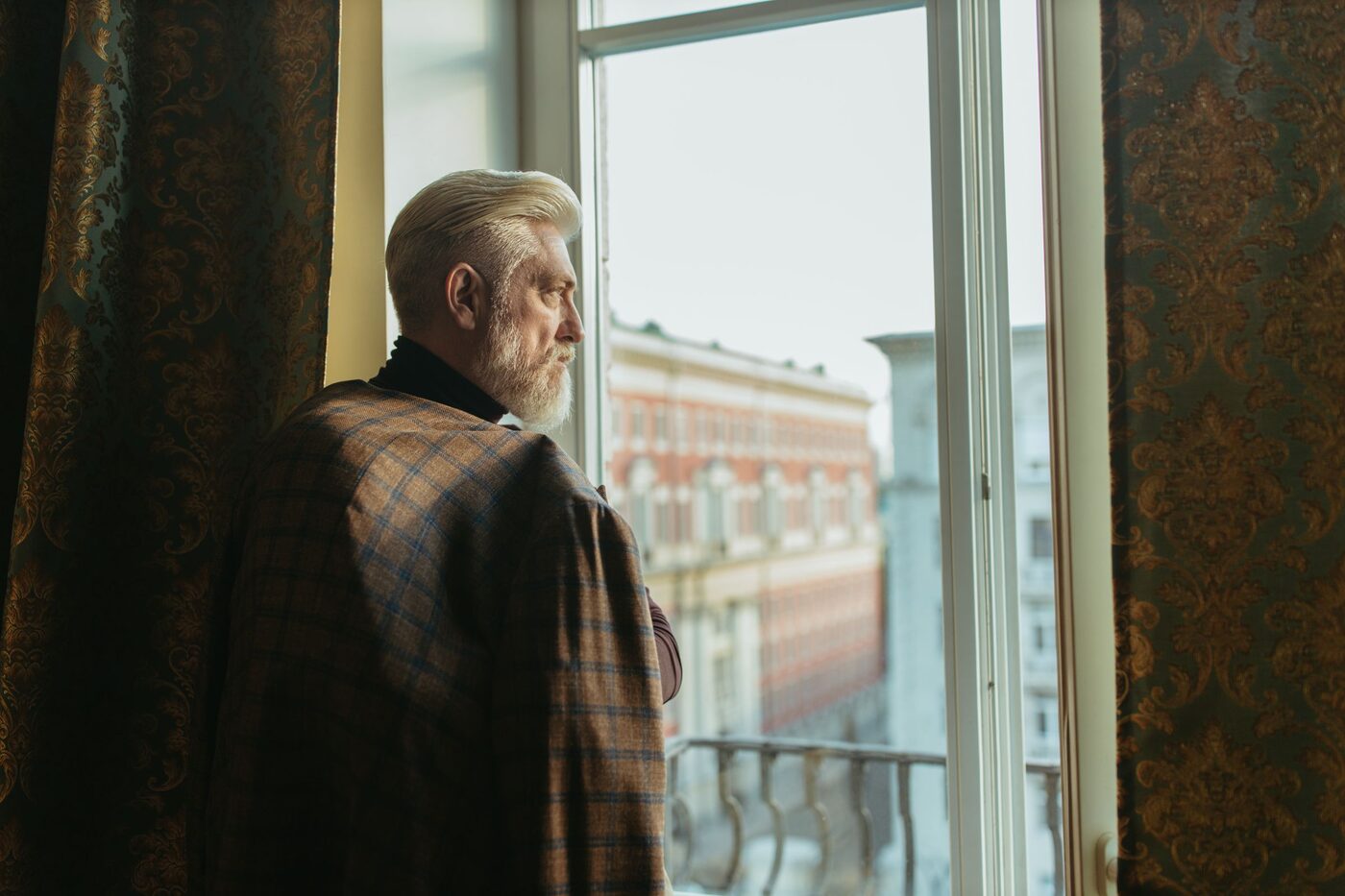 An older man standing at a tall window in soft daylight, looking out over the street below.