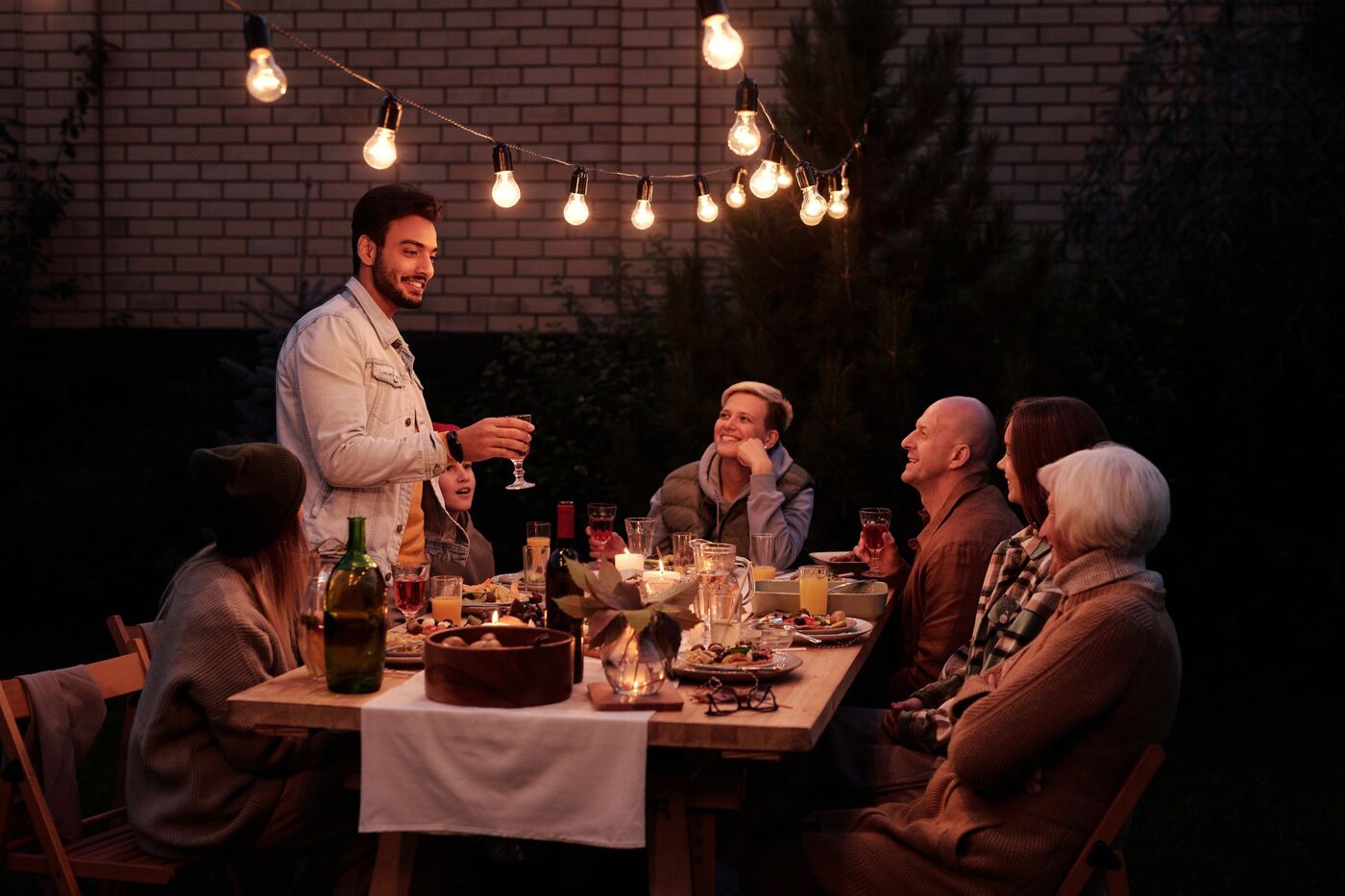 A group of friends of mixed ages eating together at a long outdoor table under strung festoon lights.